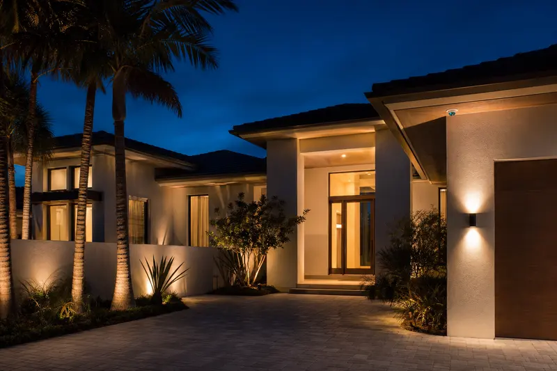 The front elevation of a coastal home at dusk, warm light in the windows, a discreet dome camera under the soffit and landscape lighting grazing the wall.